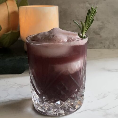 A dark red tart cherry mocktail with a sprig of rosemary poured over ice in a crystal glass