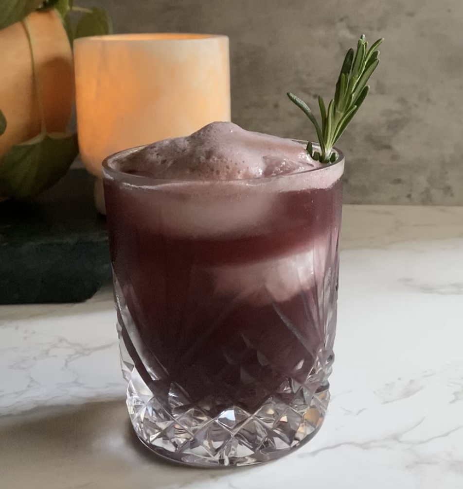 A dark red tart cherry mocktail with a sprig of rosemary poured over ice in a crystal glass