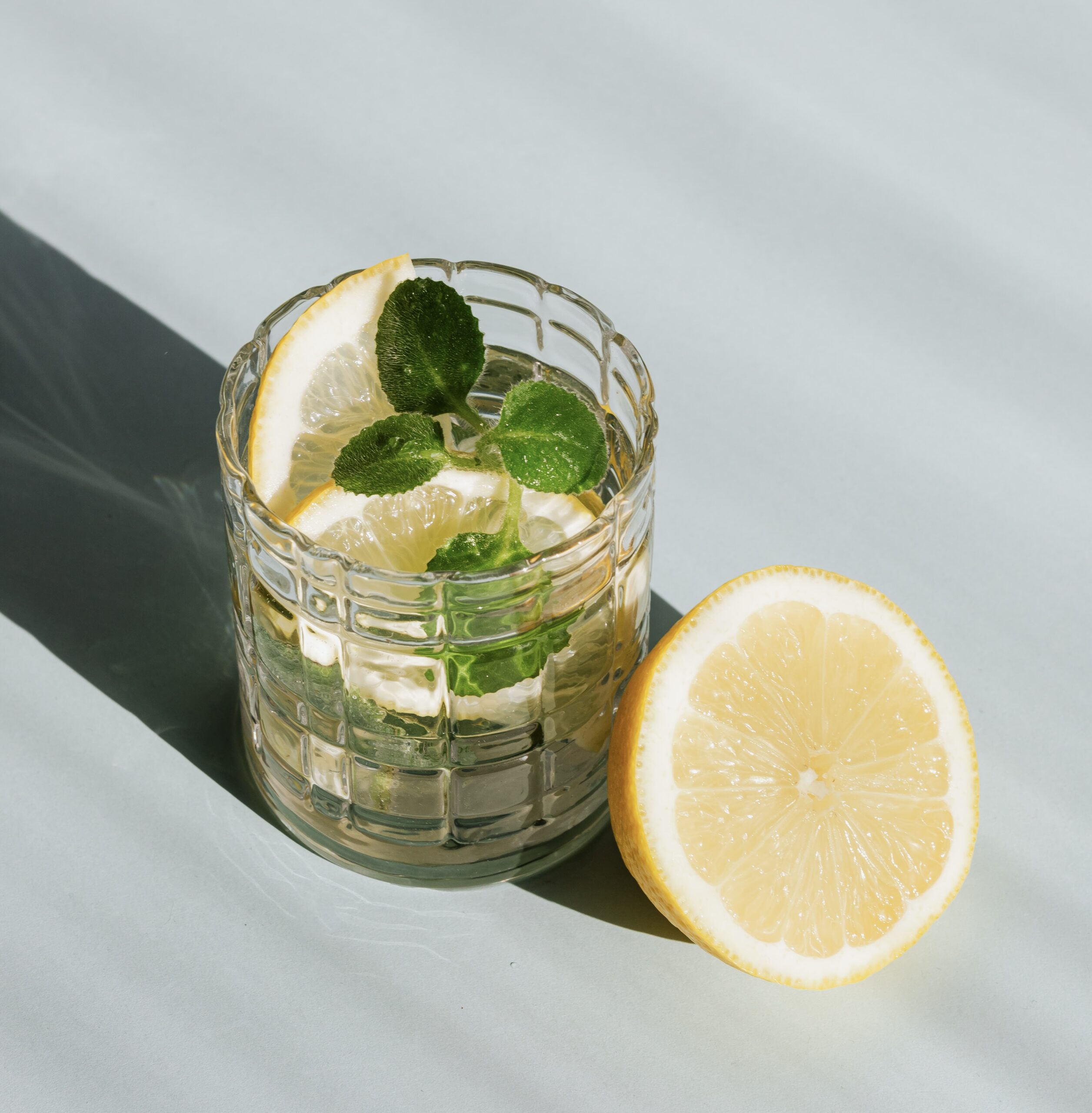 A crystal glass of water with lemon and herbs sits in the direct sunlight